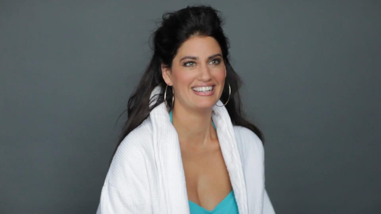 Dark-haired woman wearing a white robe and turquoise top against a gray background