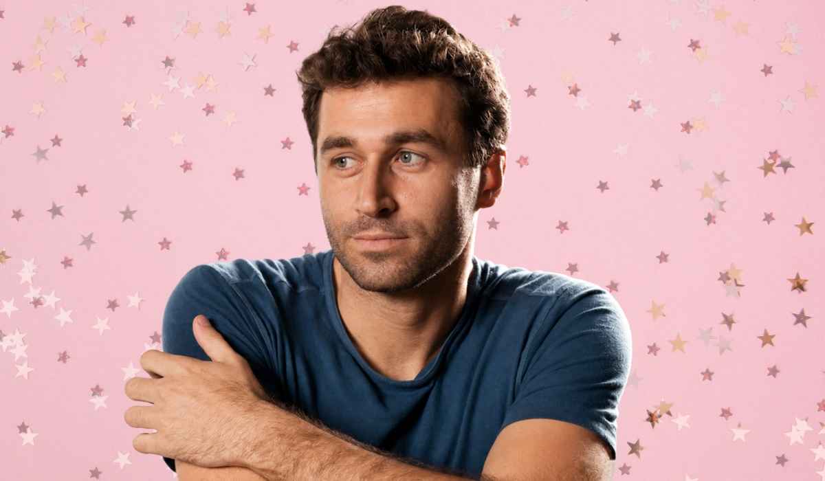 James Deen wearing a blue T-shirt with his arms crossed against a pink background