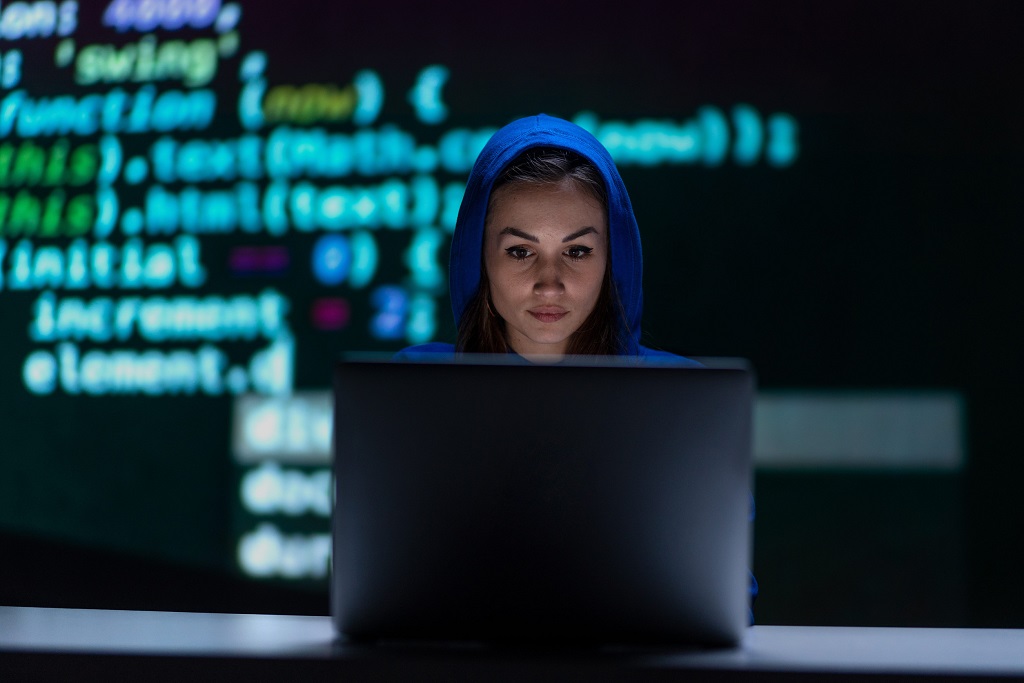 Hooded anonymous hacker woman by computer in the dark room at night, cyberwar concept.