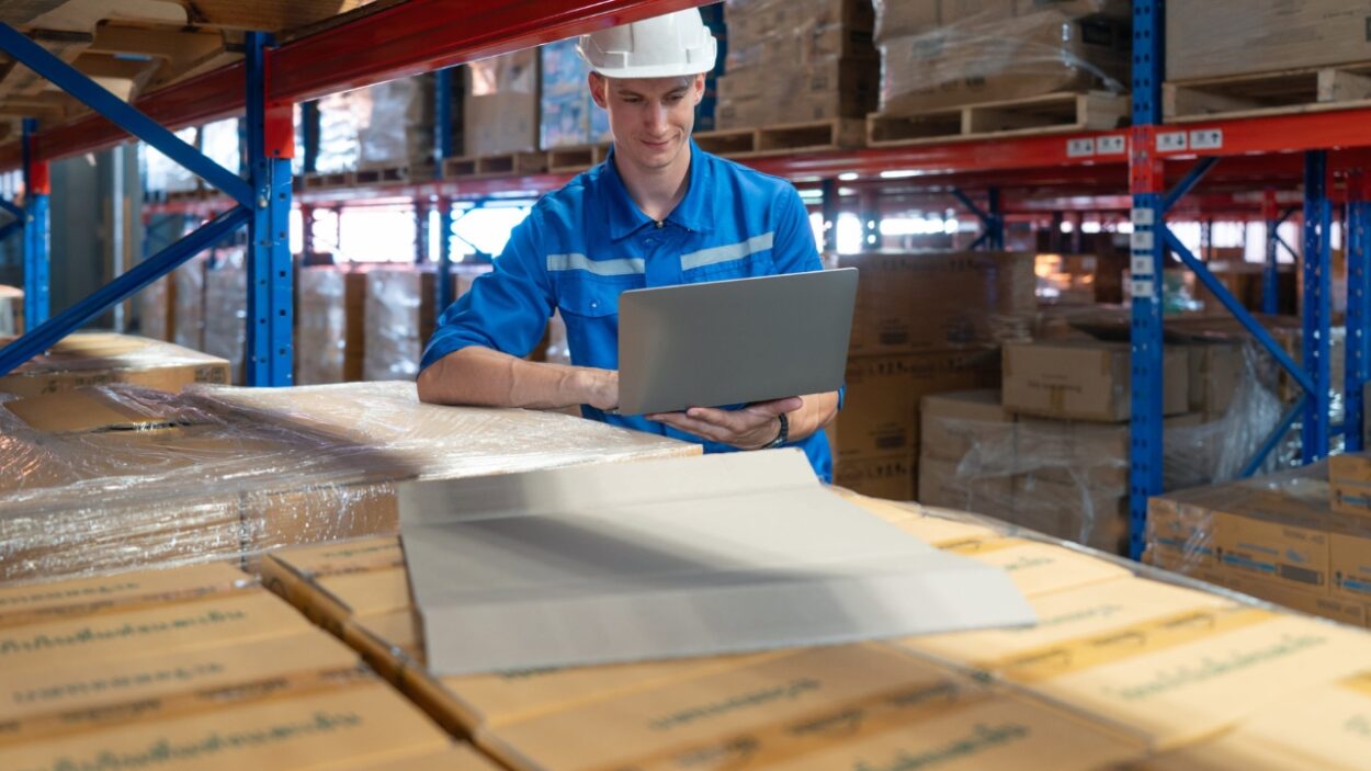 Warehouse worker uses a laptop beside stacked boxes and storage racks