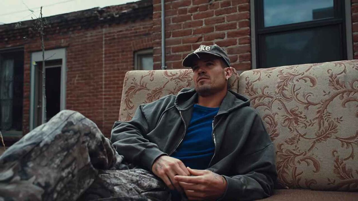 Endurance runner William Goodge sitting on a patterned couch outdoors, wearing a black cap and gray hoodie.